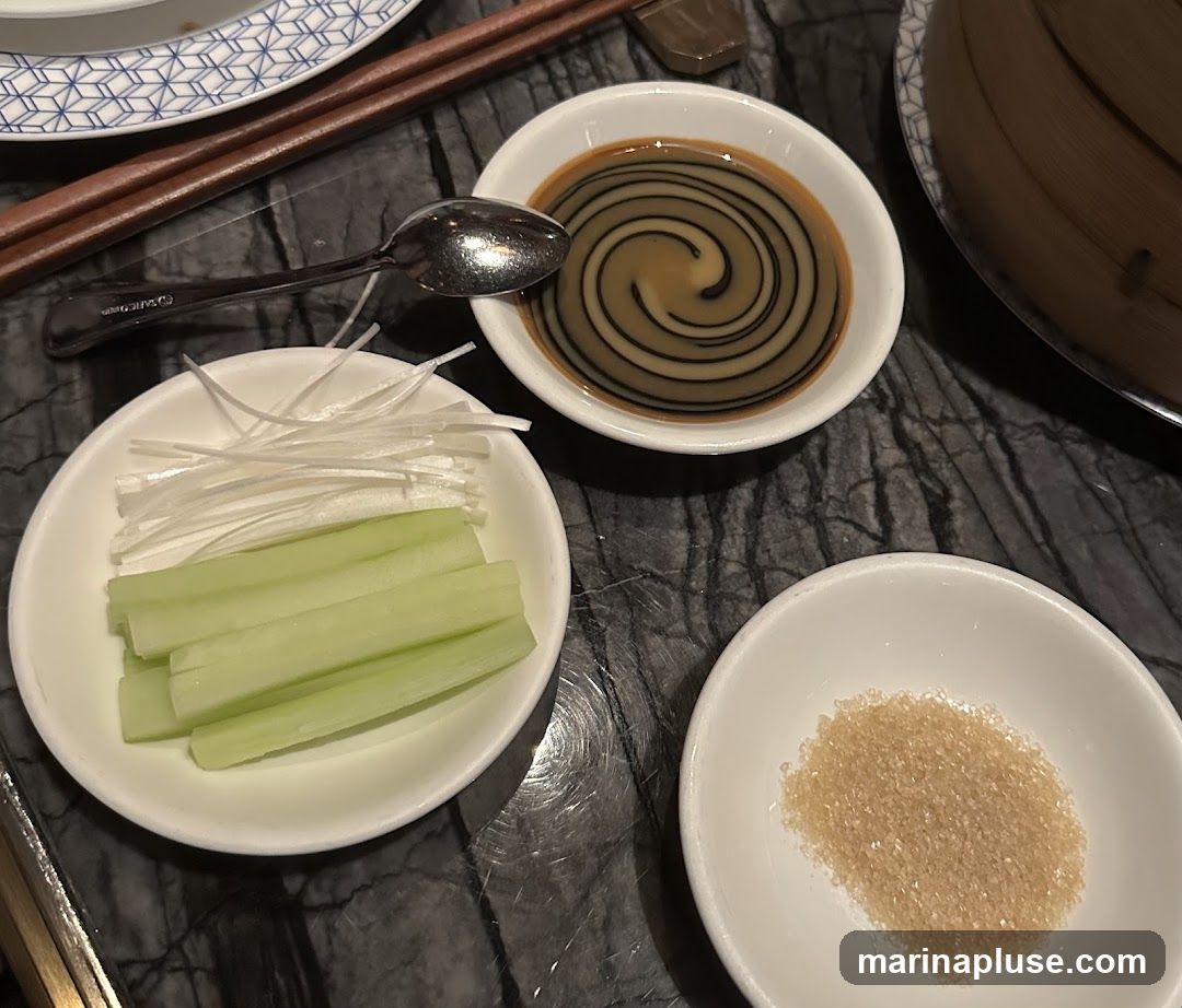 A close-up view of three small white bowls on a dark, marbled surface. The bowl in the foreground right contains a pile of light brown granulated sugar. The bowl in the foreground left holds julienned white scallions and sliced cucumber sticks. The bowl in the background contains a swirl of dark sauce, possibly hoisin or plum sauce, with a spoon resting on its edge. Chopsticks are visible in the upper left, and a bamboo steamer basket is partially visible in the upper right.
