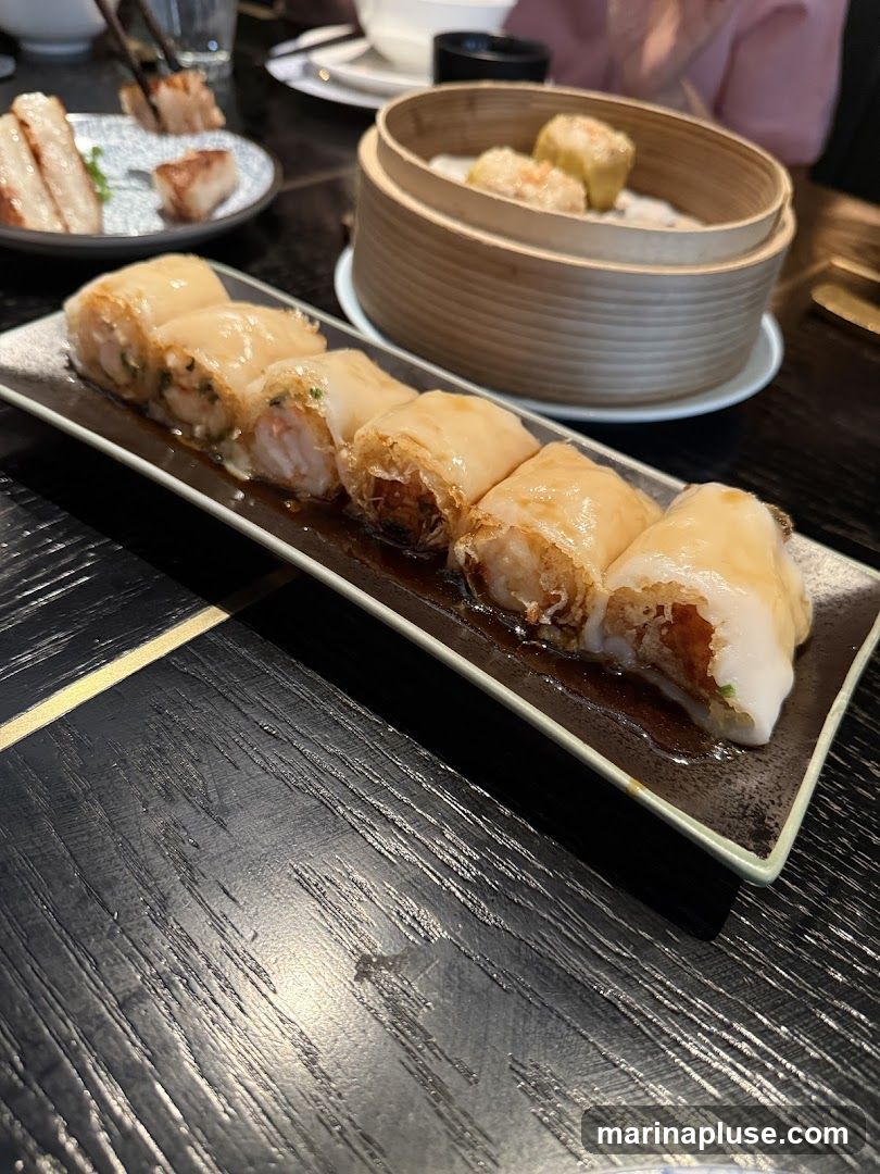 A close-up shot of a dim sum platter featuring several shrimp rice noodle rolls. In the background, a bamboo steamer basket holds more dim sum, and a plate with pork belly and other small dishes is visible. The setting appears to be a restaurant with dark wooden tables.