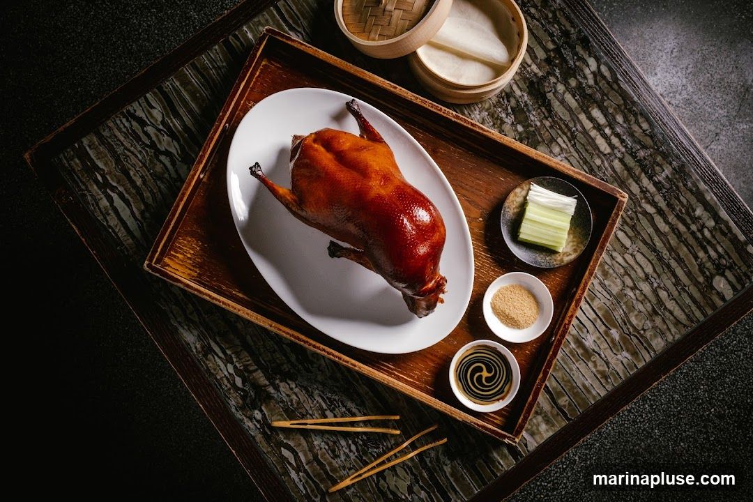 A whole roasted Peking duck is presented on a white oval platter, resting on a wooden tray. Accompanying the duck are small dishes containing sliced scallions, a pile of finely ground spice, and a swirl of dark sauce. A bamboo steamer basket filled with thin pancakes is also visible on the tray. Chopsticks lie beside the platter. The setting appears to be a table with a dark, textured surface.