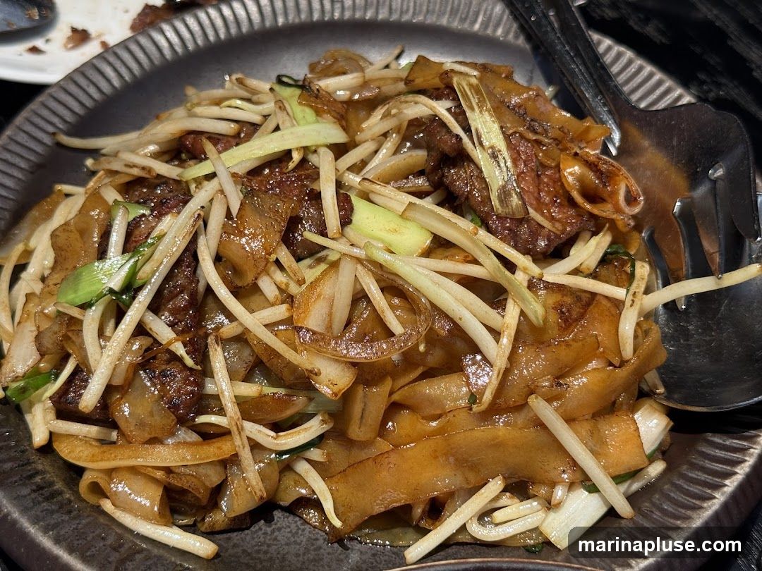 A close-up shot of beef chow fun, a popular Chinese noodle dish, served on a dark, textured plate. The dish features wide, flat rice noodles stir-fried with thinly sliced beef, bean sprouts, and green onions. The noodles are glossy and appear to be coated in a savory sauce. A fork and spoon are visible on the right side of the plate, ready to serve.