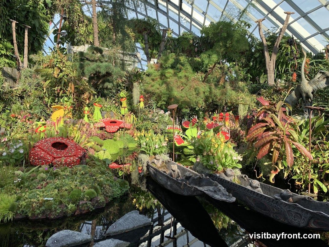A lush and vibrant display of various plants and flowers within a greenhouse. The scene features a variety of pitcher plants, some with red and yellow pitchers, and others with green foliage. There are also red, flower-like structures resembling Rafflesia flowers, and clusters of delicate white and yellow flowers. In the foreground, two intricately carved wooden canoes float on a dark body of water, reflecting the surrounding greenery. Stone sculptures of fish are partially submerged in the water. The background is filled with dense foliage, trees, and the glass ceiling of the greenhouse.