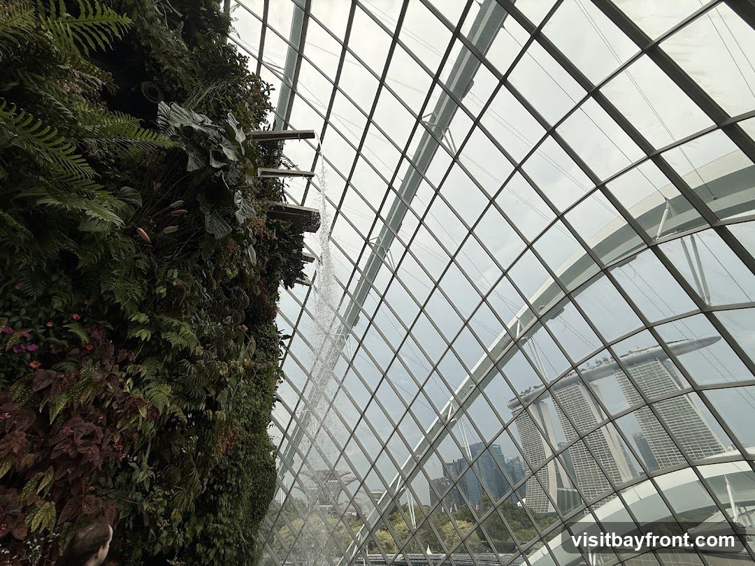 A lush vertical garden with a cascading waterfall is seen inside a geodesic dome. Through the glass ceiling, the iconic Marina Bay Sands hotel and the Singapore skyline are visible under a cloudy sky.