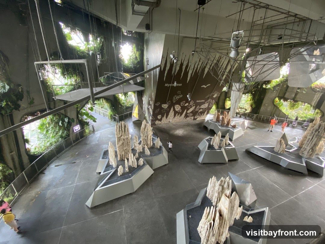An indoor exhibit featuring rock formations and lush greenery. Several displays of tall, white, crystalline rock structures are arranged on the floor. In the background, a large, dark brown wall features a graphic with text and illustrations, possibly educational. The space is designed to resemble a natural environment with hanging plants and a walkway visible in the distance. A few visitors are seen walking through the exhibit.