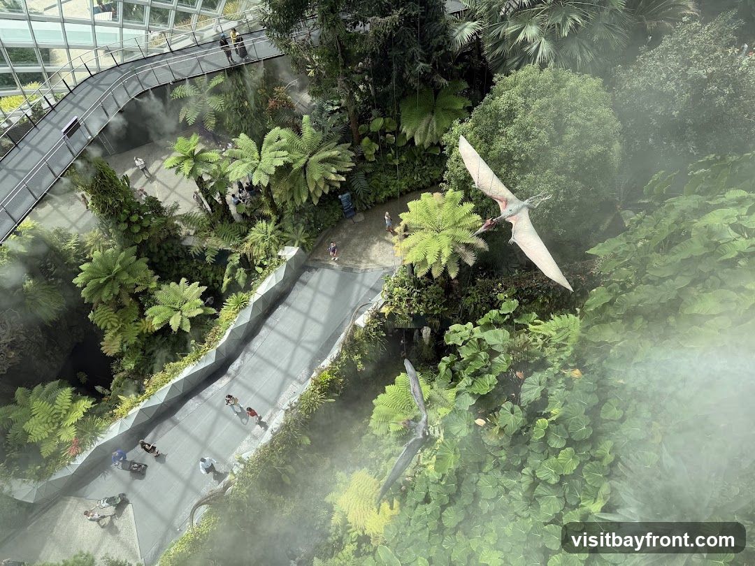 An aerial view of a lush indoor botanical garden, possibly a conservatory or greenhouse. A walkway winds through dense foliage, with various types of ferns and trees. Mist is being sprayed, creating a hazy atmosphere. Several models of pterodactyls are suspended in the air, adding a prehistoric element to the scene. People are visible walking along the pathways. The structure's glass ceiling is partially visible at the top.