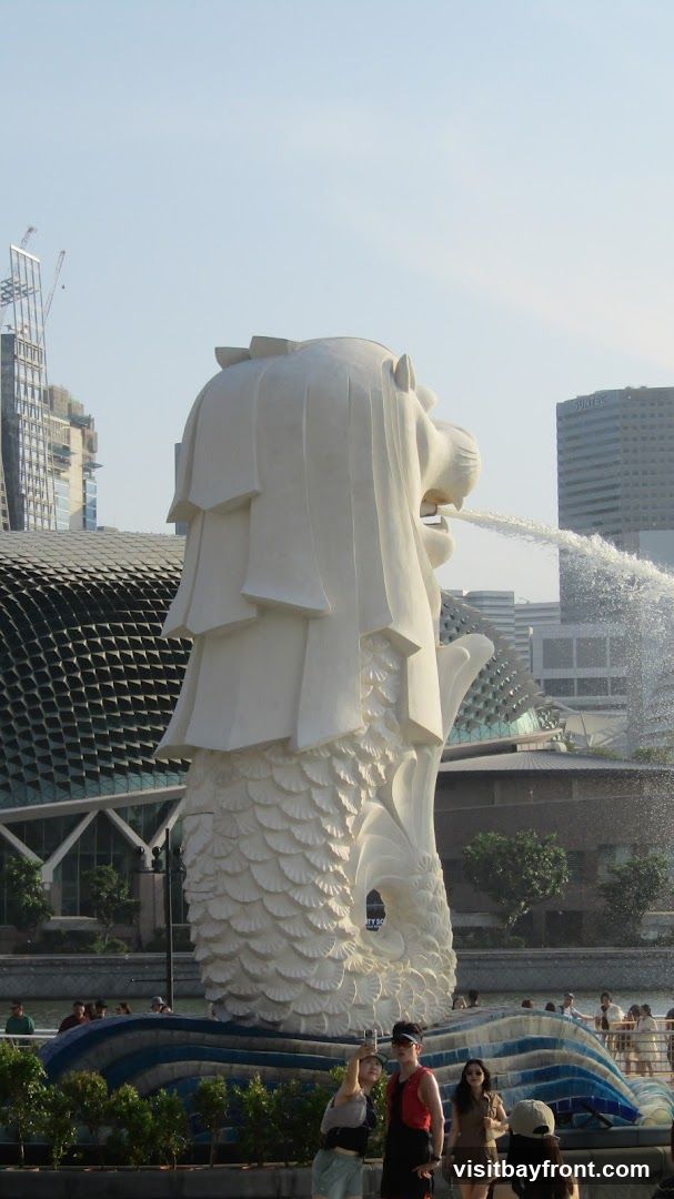 A large white statue of a mythical creature with a lion's head and a fish's body is spouting water into a pool. In the background, there are modern buildings and a distinctive domed structure. Several people are visible in the foreground and around the pool, some taking pictures.