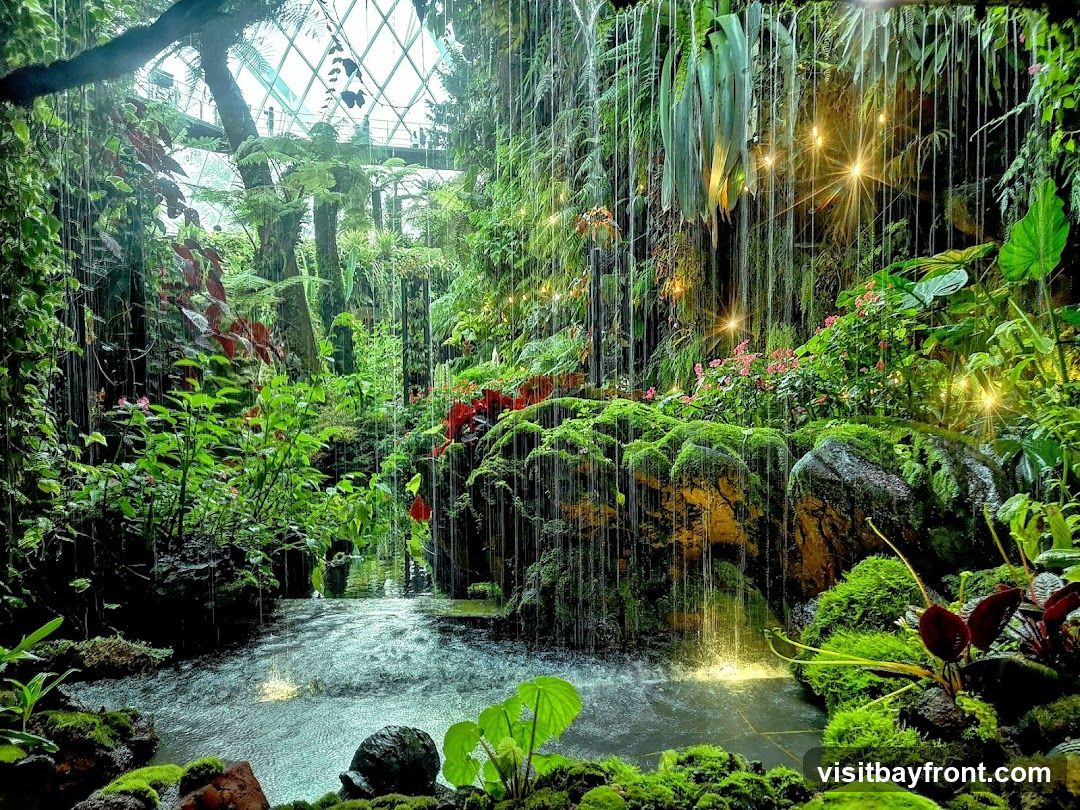 A lush indoor garden with a cascading waterfall, moss-covered rocks, and abundant greenery. String lights are visible, adding a warm glow to the scene. The environment appears to be enclosed by a geodesic dome structure, suggesting a botanical garden or conservatory.
