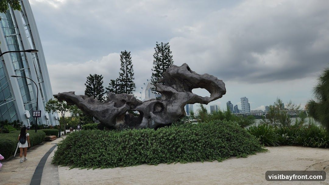 A large, weathered rock sculpture is displayed in a lush garden setting. The sculpture has several openings and an organic, abstract shape. Behind it, tall evergreen trees stand against a cloudy sky. In the distance, a Ferris wheel and modern city buildings are visible, along with a body of water and a bridge. A pathway curves through the garden, and a person is walking away from the camera on the left side. The architecture of a large, glass-paneled building is visible on the far left.