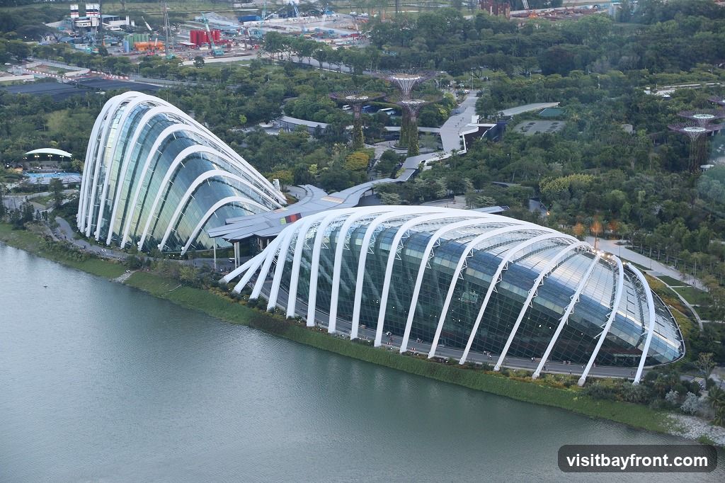 An aerial view of the large, curved glass conservatories at Gardens by the Bay, Singapore, situated next to a body of water. The structures feature a distinctive white lattice framework supporting the glass panels. In the background, lush greenery and the iconic Supertree Grove are visible, along with some construction activity in the distance.