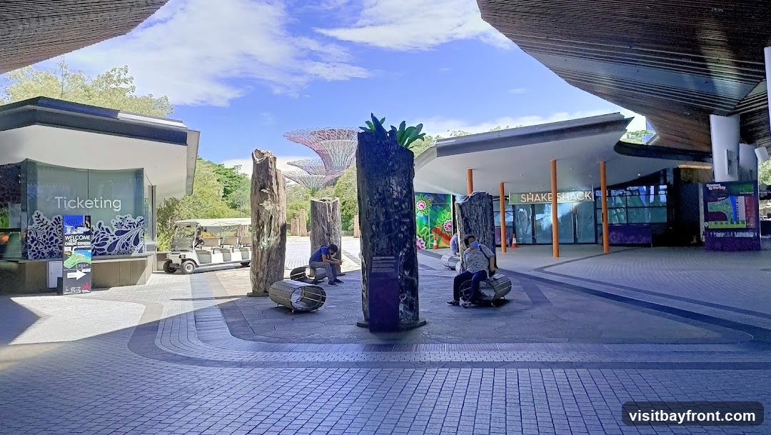 An outdoor plaza with a ticketing booth on the left and a Shake Shack restaurant on the right. Several people are seated on benches in the center of the plaza, which is decorated with large wooden pillars. In the background, the iconic Supertrees of Gardens by the Bay are visible. A golf cart is parked near the ticketing booth.