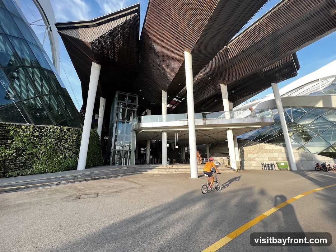 A wide shot of a modern building with a distinctive architectural design. The building features a large, cantilevered roof structure with wooden slats, supported by white columns. To the left, a glass facade is covered with a lush green wall of plants. An elevator is visible in the center, leading to an elevated walkway. In the foreground, a person rides a bicycle on a paved area with a yellow line marking. The sky is blue with some clouds.