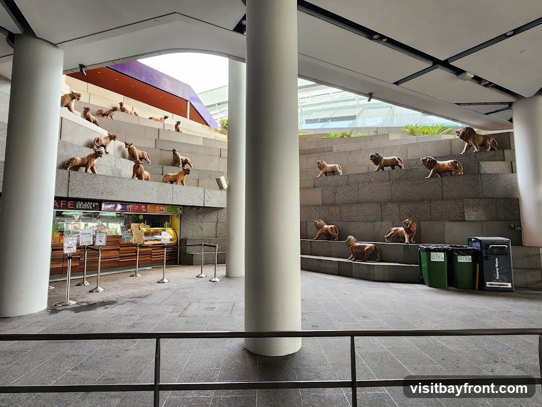 A wide shot of an outdoor seating area with tiered stone steps. Numerous sculptures of lions are placed on the steps, facing in various directions. Below the steps, a cafe counter is visible with display cases of food items. The cafe has signage that reads 'CAFE ICE CREAM & SNACKS'. Several stanchions and a trash bin are also present in the scene.