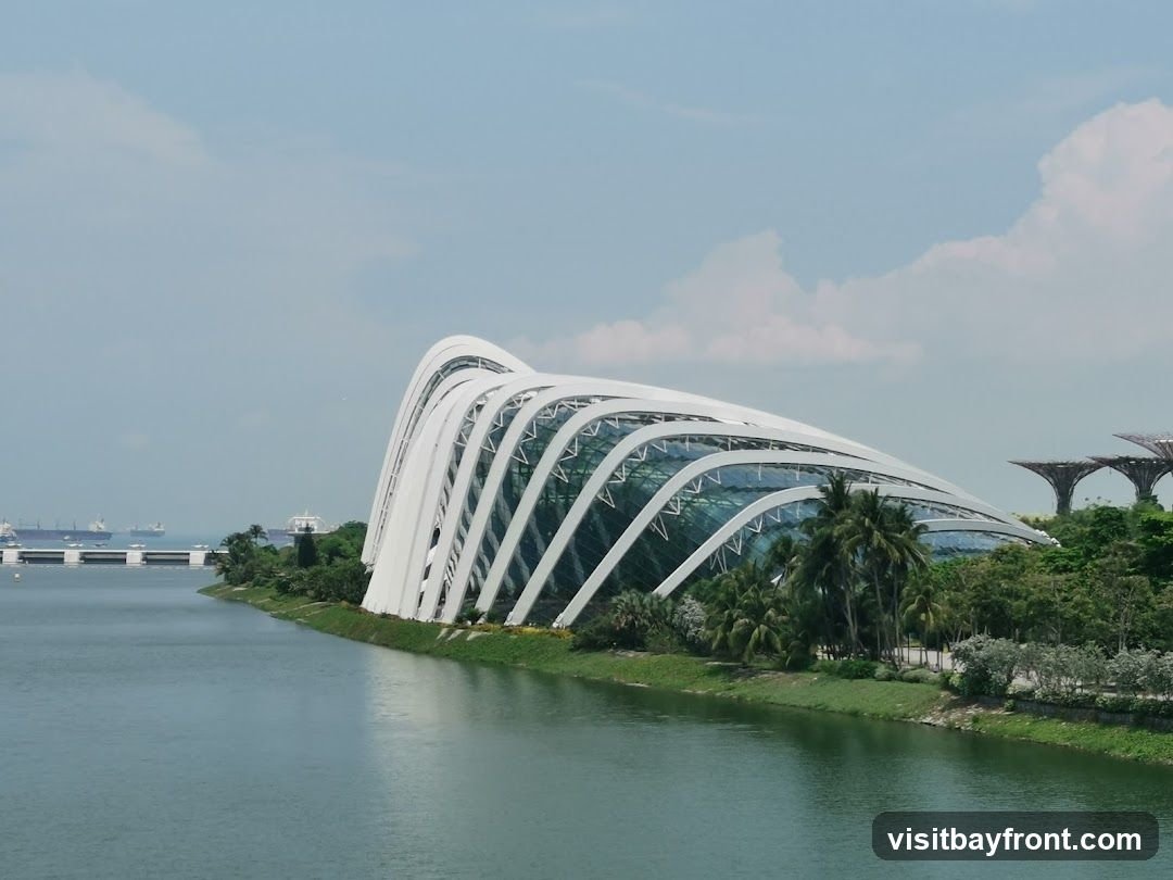 A wide shot of the iconic Flower Dome and Cloud Forest conservatories at Gardens by the Bay, Singapore. The futuristic, curved structures with white lattice frameworks and glass panels are set against a backdrop of lush greenery and a calm body of water. In the distance, several cargo ships are visible on the horizon, and the Supertree Grove can be seen to the right. The sky is partly cloudy with a light blue hue.