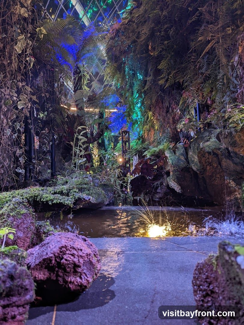A lush indoor garden with a variety of plants, including ferns and vines, illuminated by colorful blue and green lights. A small water feature with a fountain creates ripples and splashes in a shallow pool. Large, textured rocks covered in moss are visible in the foreground and around the water. The scene is enclosed by a glass ceiling structure.