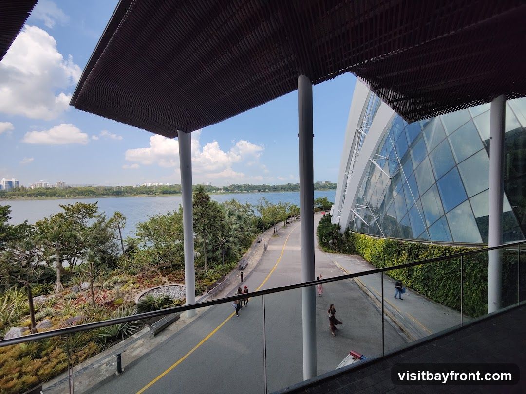 A view of the exterior of a large, modern conservatory with a glass facade, surrounded by lush greenery and a body of water. People are seen walking along a path outside the structure. The sky is blue with scattered clouds.