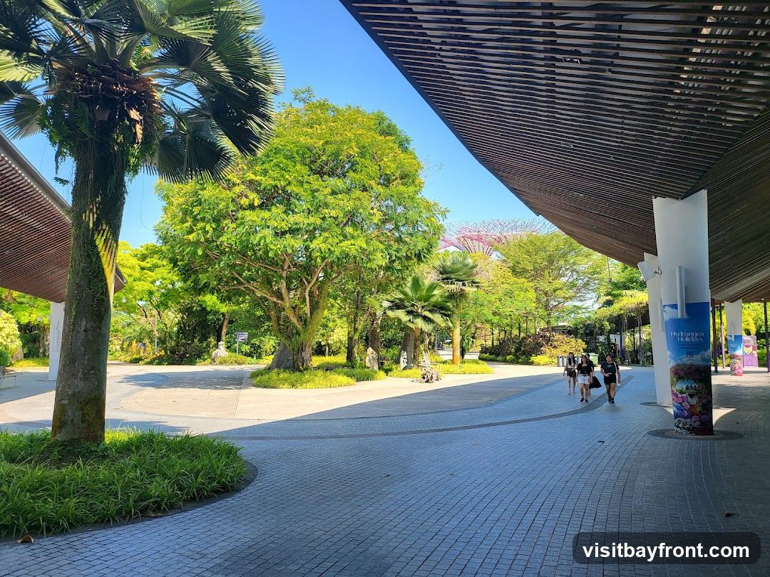 A paved walkway under a modern, slatted roof structure leads through a lush green park. Palm trees and other foliage are abundant, with a glimpse of the iconic Supertree Grove in the distance. Several people are walking along the path. A vertical banner on a pillar advertises "Hortsea Holidays" with floral imagery.