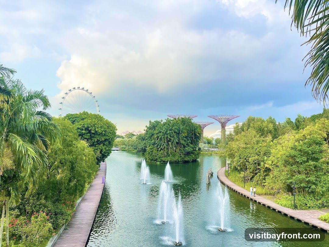 A scenic view of Gardens by the Bay in Singapore, featuring a tranquil body of water with fountains, lush greenery, and iconic Supertree Grove structures in the background. A Ferris wheel is also visible on the horizon. Wooden boardwalks line the water, with a few people strolling along them. The sky is partly cloudy, with soft light illuminating the landscape.