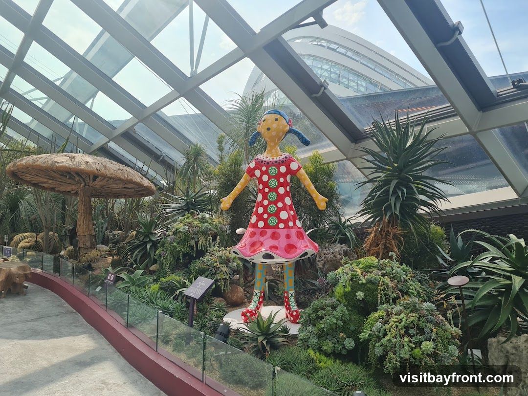 A whimsical sculpture of a girl in a polka-dot dress stands amidst a lush display of succulents and cacti within a glass-domed conservatory. A large mushroom-shaped structure is visible to the left, and the distinctive architecture of the conservatory's roof and a distant building are seen overhead.