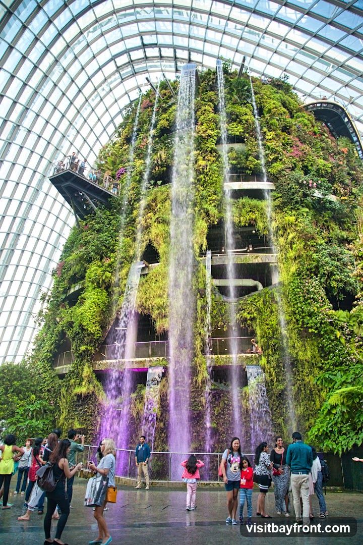 A large indoor garden with a massive, multi-tiered waterfall cascading down a lush, plant-covered structure. People are visible on walkways and on the ground level, observing the attraction. The space is enclosed by a curved glass ceiling.