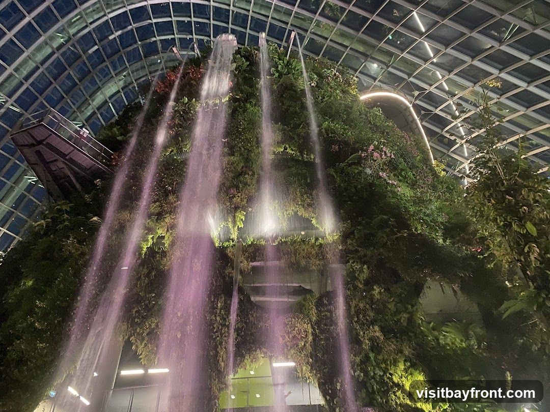 A view looking up at a large, multi-tiered indoor waterfall cascading down a lush, green, plant-covered structure. The waterfall is illuminated with purple and white lights, creating a dramatic effect. Above, a glass and metal grid ceiling is visible. A walkway or platform can be seen on the left side of the structure. The overall scene depicts a vibrant, artificial natural environment within a large building.