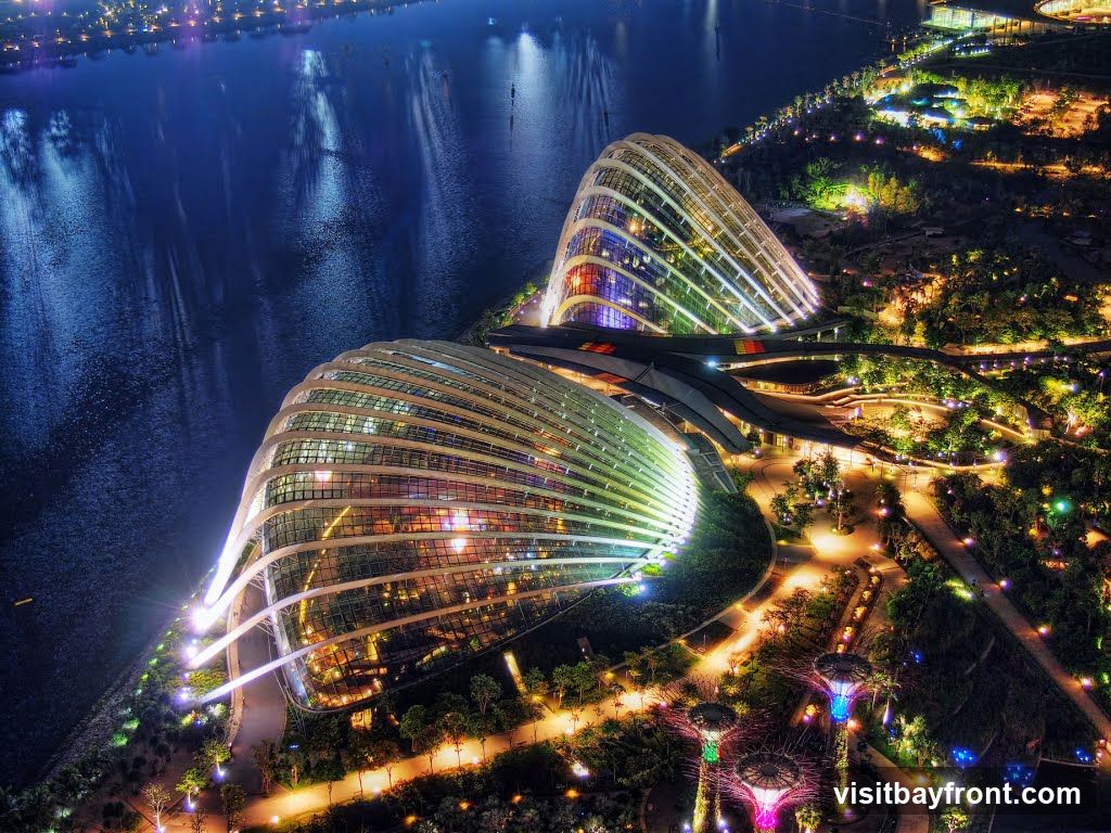 An aerial nighttime view of Gardens by the Bay in Singapore, showcasing the illuminated Supertree Grove and the iconic Cloud Forest and Flower Dome conservatories. The structures are lit with vibrant colors, reflecting on the adjacent water body. The surrounding gardens are also illuminated with pathways and decorative lights, creating a magical landscape.