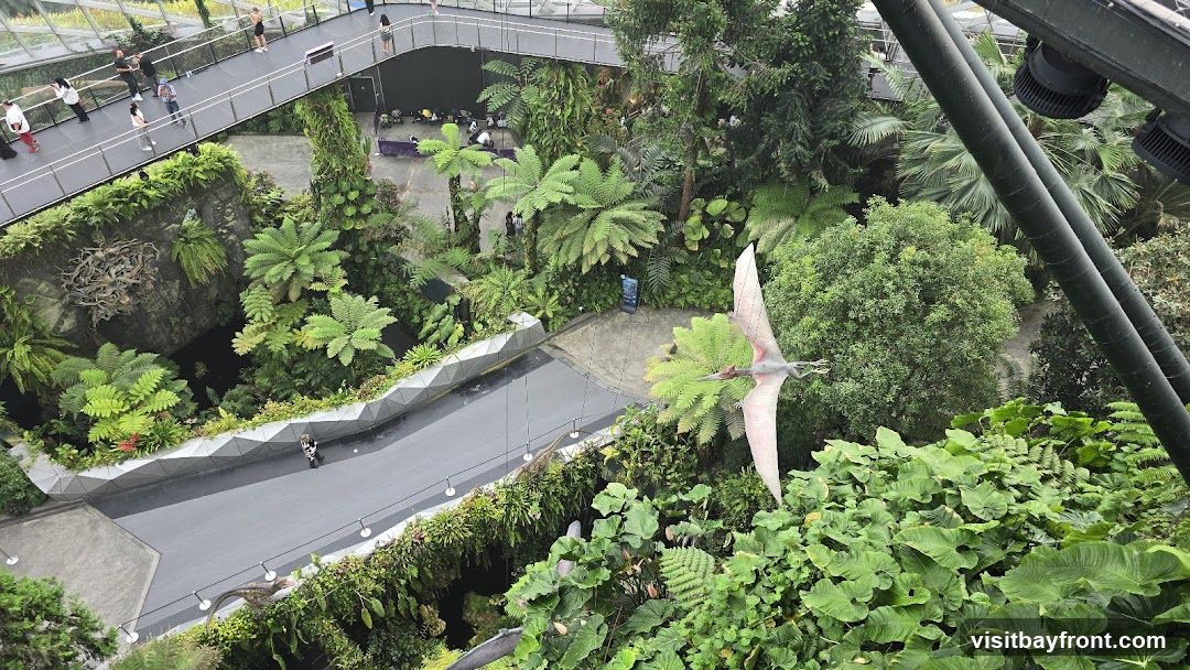 An aerial view of a lush indoor botanical garden, possibly a conservatory or greenhouse. Elevated walkways with visitors are visible, winding through dense foliage. Several large, realistic dinosaur models, including a pterodactyl in flight and a terrestrial dinosaur, are placed among the plants. The scene is characterized by abundant greenery, diverse plant species, and artificial lighting fixtures overhead.