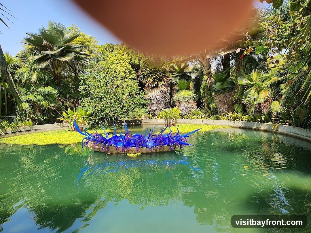 A vibrant blue glass sculpture floats in a tranquil pond surrounded by lush tropical foliage. The sculpture, resembling a cluster of spiky, organic forms, is positioned on a raft covered in small green plants. The water is a clear, emerald green, reflecting the surrounding greenery and the bright blue sky. Palm trees and other exotic plants create a dense, verdant backdrop, suggesting a botanical garden or a serene outdoor oasis. A blurred orange shape, possibly a finger, is visible in the upper portion of the image.