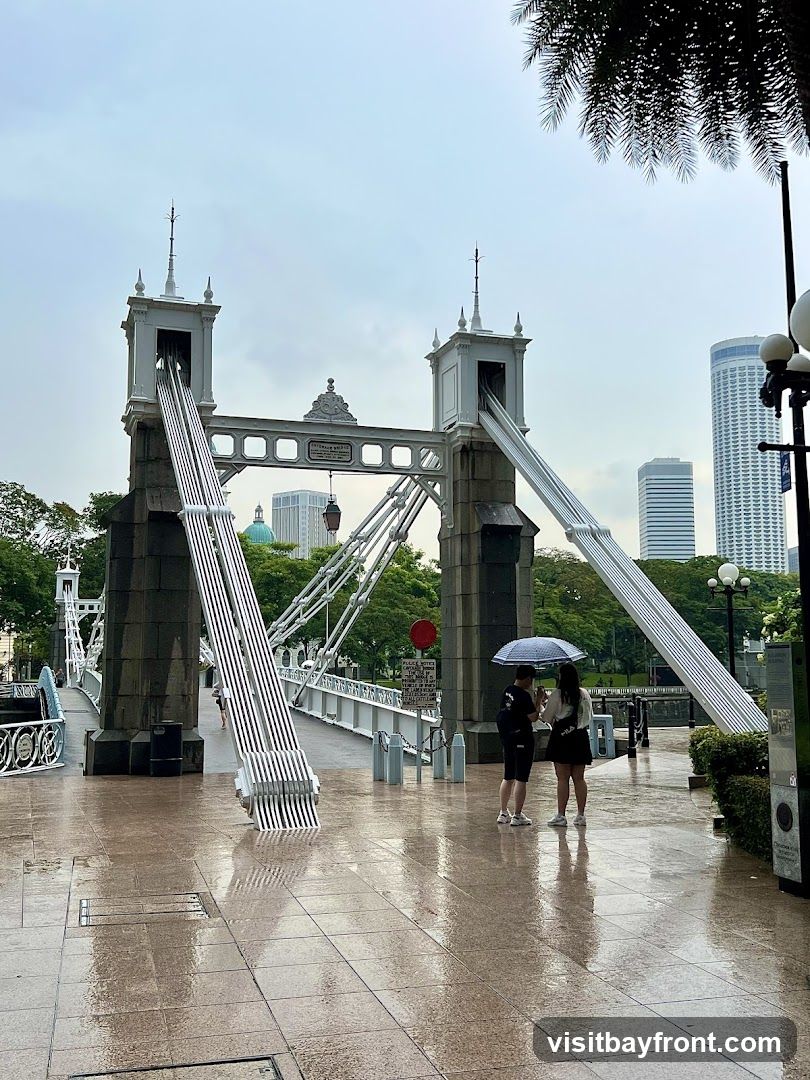 Two people stand under an umbrella on a wet bridge, with modern buildings in the background. The bridge has distinctive white towers and suspension cables. The ground is wet and reflective, suggesting it has been raining.