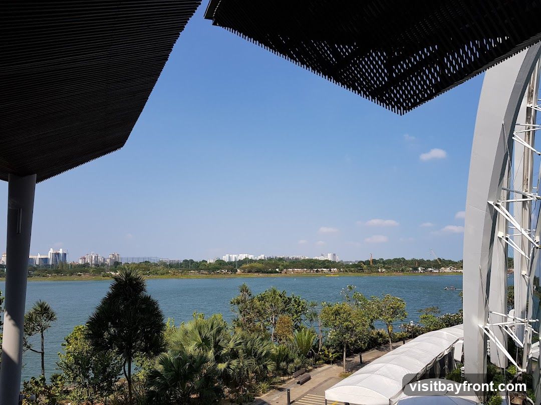 A scenic view of a calm body of water with a distant city skyline under a clear blue sky. Lush green trees and tropical plants line the waterfront. In the foreground, modern architectural elements, including a dark, slatted overhang and a white, arched structure with metal supports, frame the scene. White tents or awnings are visible along the edge of the water.