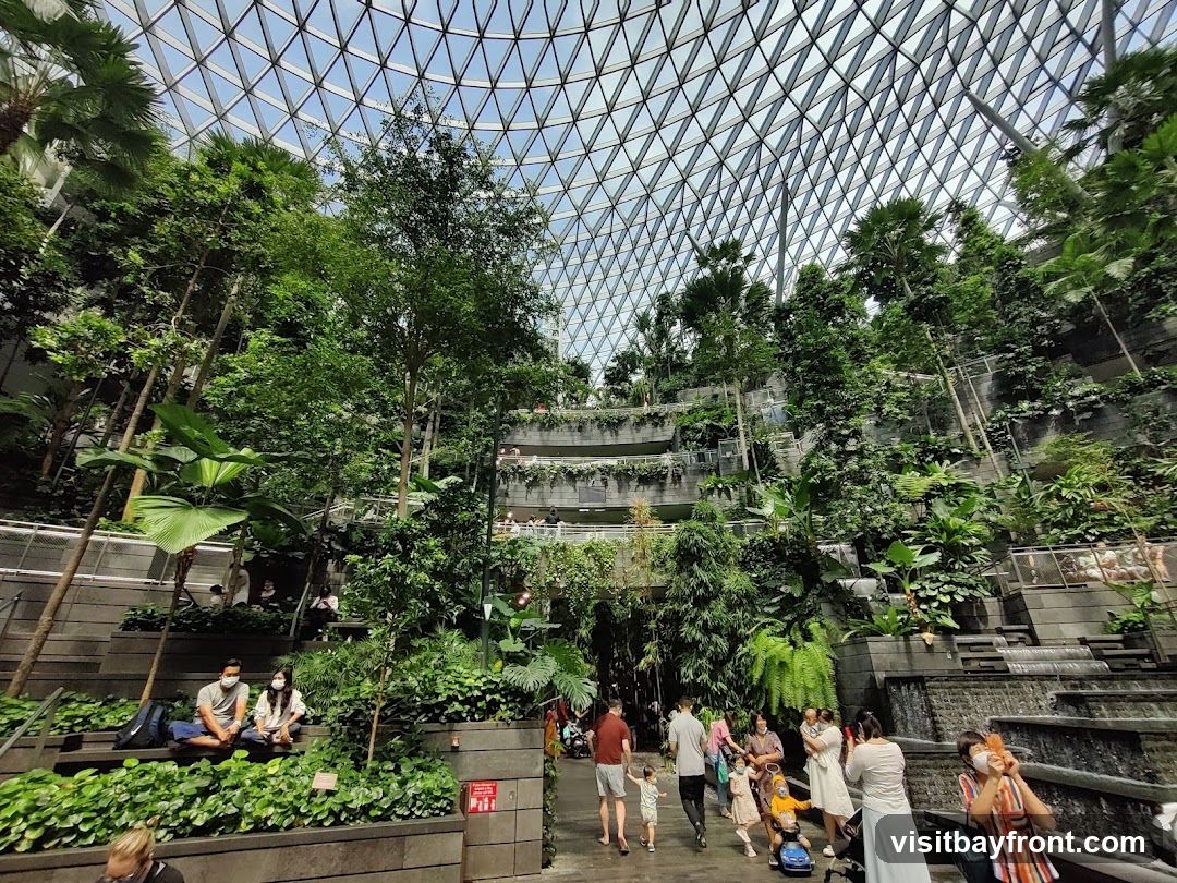 A lush indoor garden with tiered levels of plants and trees, enclosed by a geodesic dome roof. People are seen walking and sitting, enjoying the greenery. A multi-tiered waterfall cascades down the side.