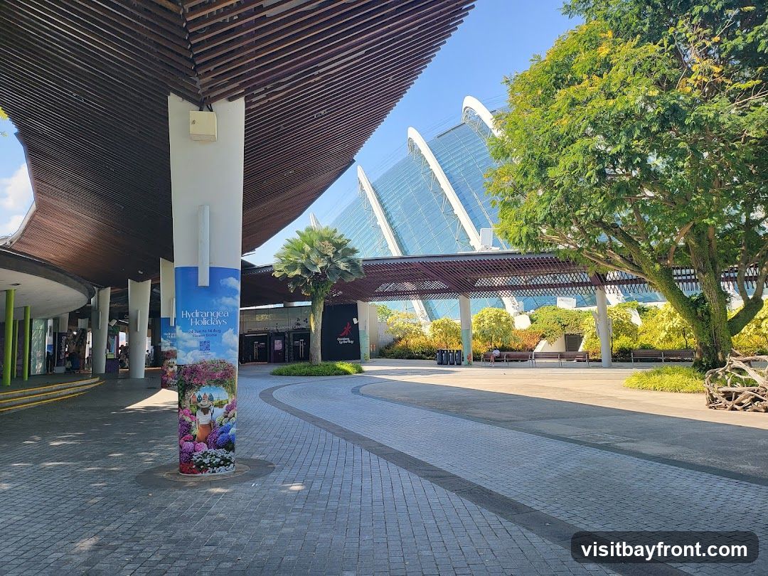 An outdoor walkway leads towards the entrance of a large, modern conservatory with a distinctive glass and steel structure. The walkway is paved with interlocking stones and lined with trees and benches. A large pillar in the foreground displays a colorful advertisement for "Hydrangea Holidays." The overall scene suggests a public garden or park on a sunny day.