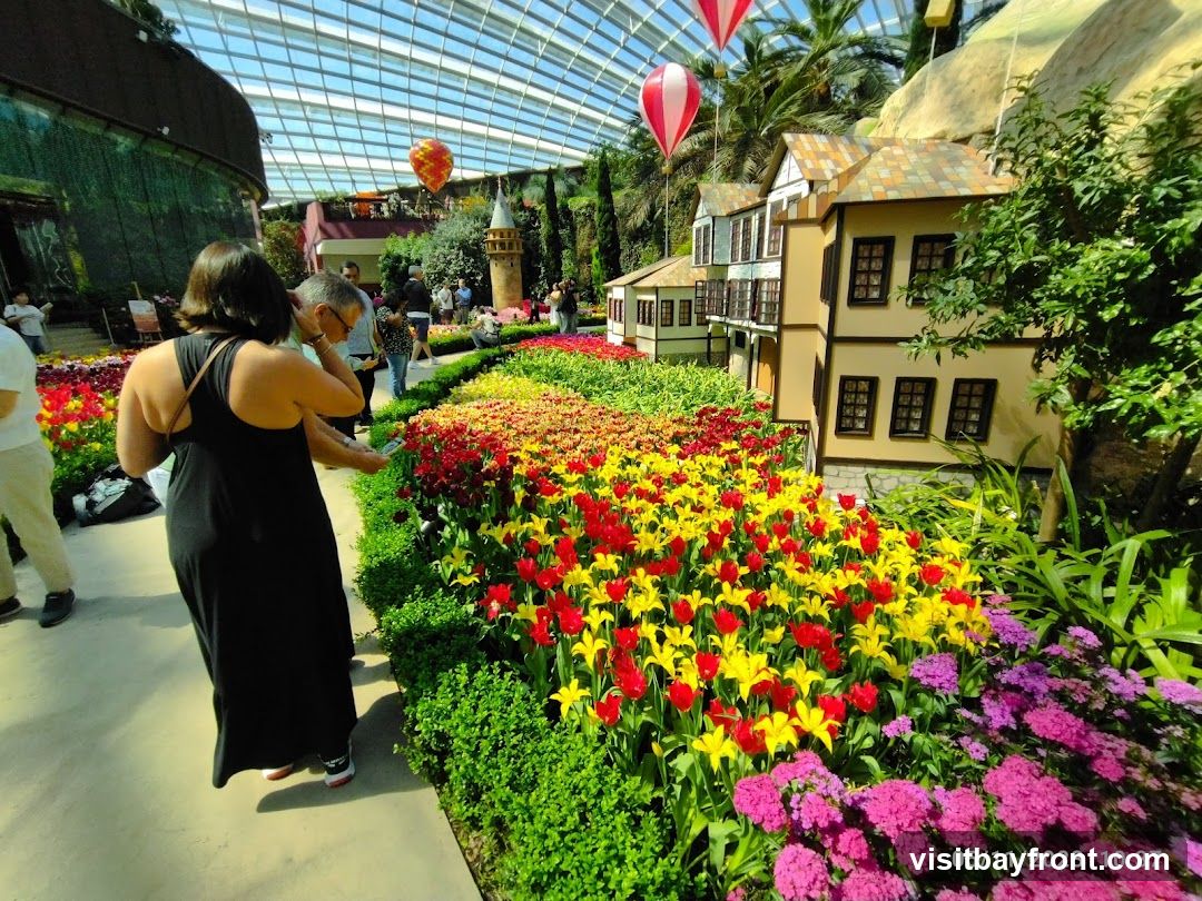 A vibrant display of red and yellow tulips and pink flowers is arranged in front of miniature houses and a castle tower. Hot air balloons are suspended in the background under a glass ceiling. Several people are walking through the exhibit.