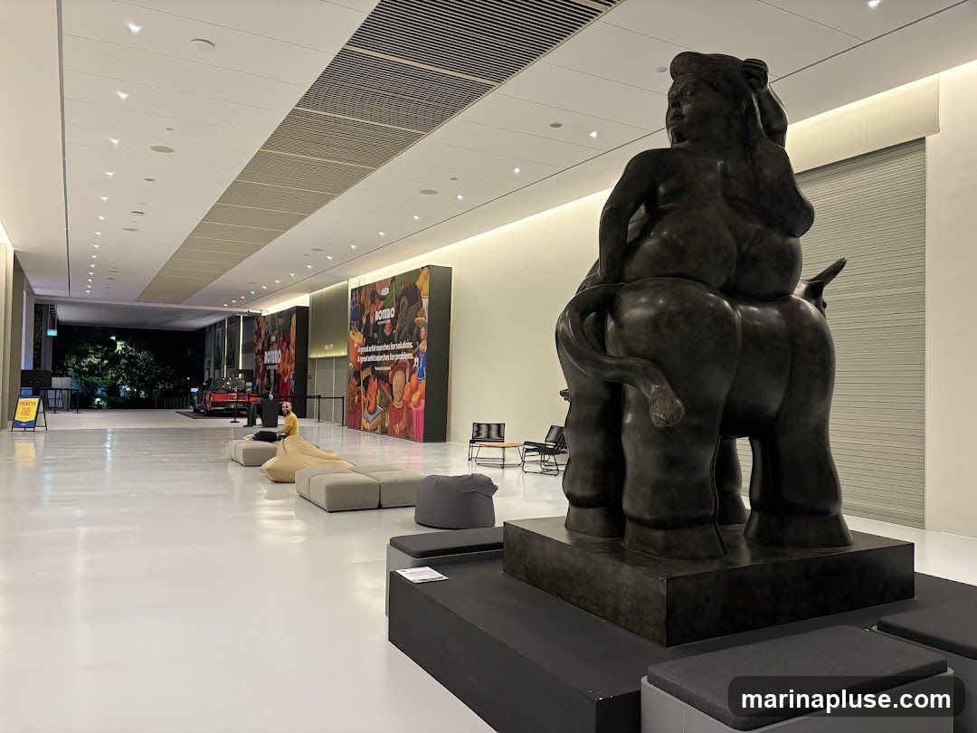 A large bronze sculpture of a seated figure on an animal is displayed in a modern, well-lit atrium. The sculpture is the focal point of the image. In the background, there are large colorful posters with text, seating areas with beanbag chairs and benches, and a red car visible through an open doorway. The space appears to be an exhibition hall or museum lobby.