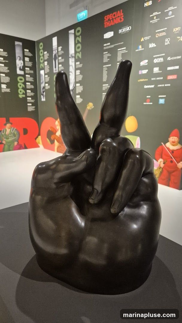 A bronze sculpture of a hand forming a peace sign is displayed in front of a wall with text and logos. The wall features sections labeled