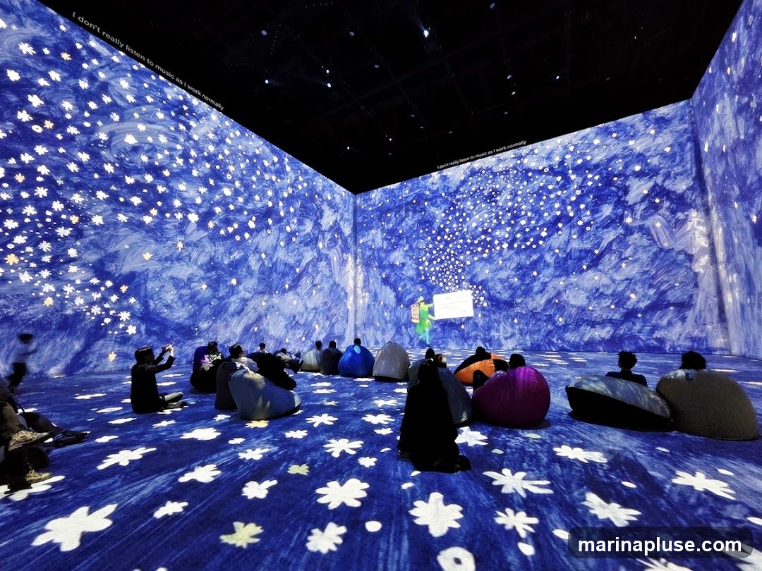 An immersive art installation with blue and white starry projections covering the walls and floor. Several people are seated on beanbag chairs, some taking photos with their phones. A figure in green stands near a projection of text.