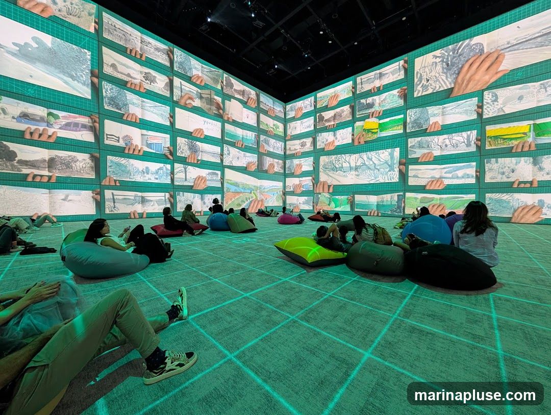People are sitting on beanbag chairs in a room with walls covered in a large projection. The projection displays a grid of images, each showing a page from a sketchbook with drawings of landscapes and hands holding the pages. The floor is also projected with a grid pattern.