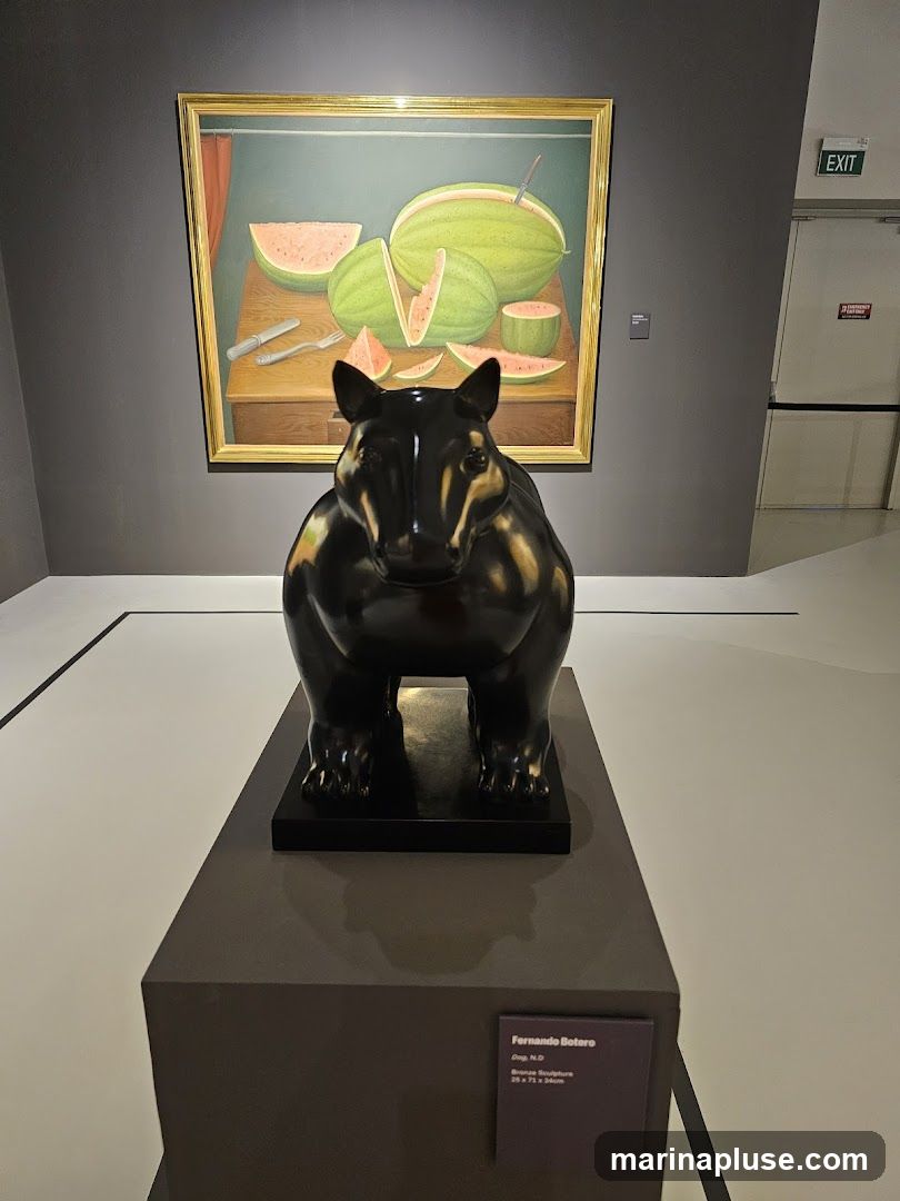 A dark, stylized sculpture of an animal stands on a pedestal in front of a framed painting of watermelons. The painting depicts several whole and sliced watermelons on a wooden table, with a knife and fork. An exit sign is visible on the wall in the background. A plaque below the sculpture identifies it as "Fernando Botero, Dog, N.D., Bronze Sculpture, 26 x 71 x 34cm.".