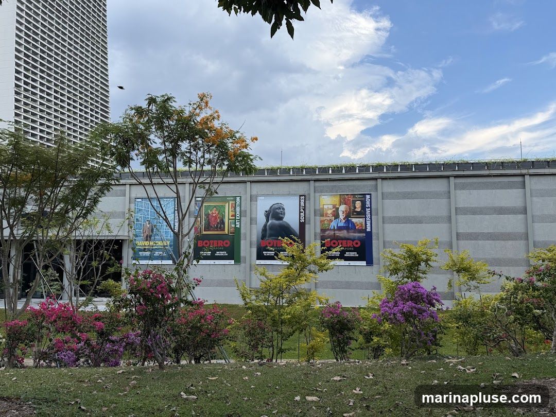 A row of large posters advertising art exhibitions is displayed on the exterior wall of a building. The posters feature artwork and portraits, with titles such as "DAVID HOCKNEY IMMERSIVE SHOW" and "BOTERO ART EXHIBITION". Lush greenery and flowering bushes are in the foreground, with a tall modern building visible in the background under a cloudy sky.