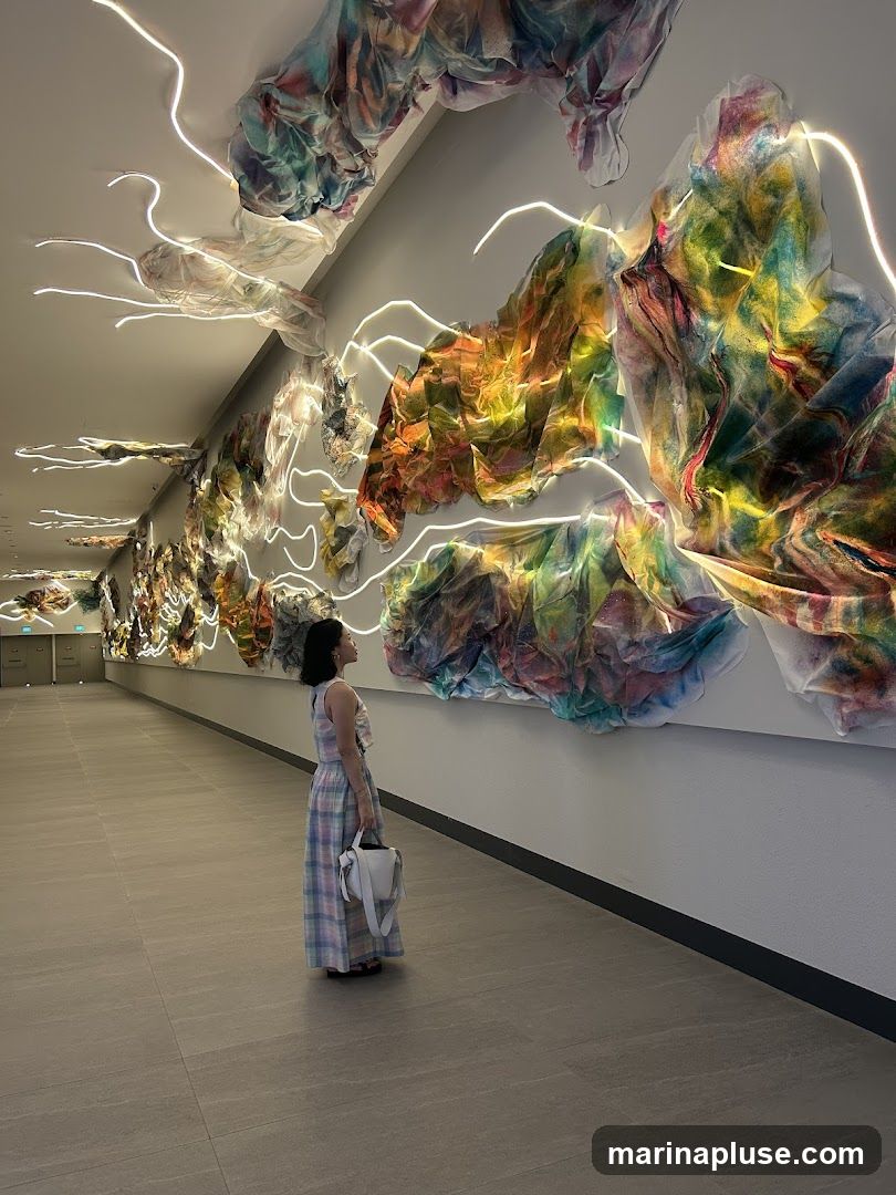 A woman in a plaid dress stands in a hallway, looking at a large, colorful art installation on the wall. The installation consists of fabric-like elements in vibrant hues of pink, yellow, green, and blue, illuminated by white LED lights that snake around the pieces. The hallway has a tiled floor and a white wall.