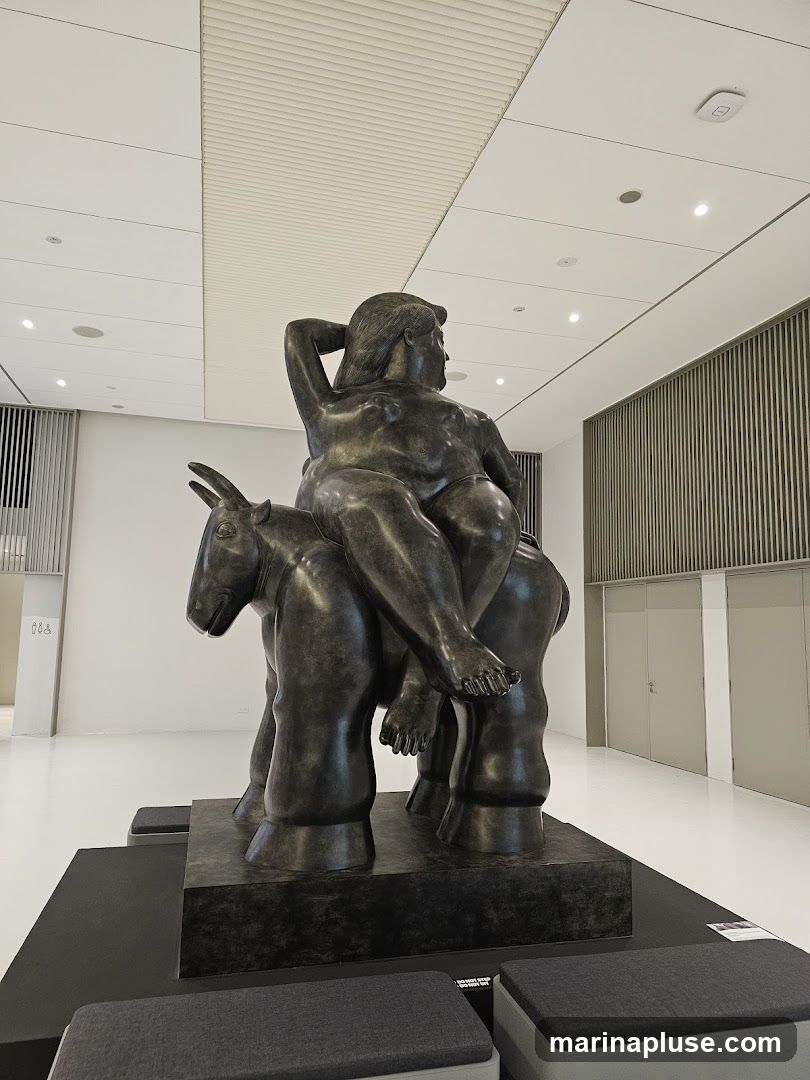 A large bronze sculpture of a woman with a full figure is depicted riding on the back of a donkey. The sculpture is placed on a dark pedestal in a well-lit, modern interior space with a white floor and ceiling. The walls are adorned with vertical slats.