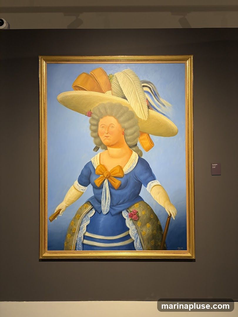 A painting of a woman with a large, ornate hat and a blue dress. She is holding a fan in one hand and a cane in the other. The painting is framed in gold and displayed on a dark gray wall. A small plaque is visible to the right of the painting.