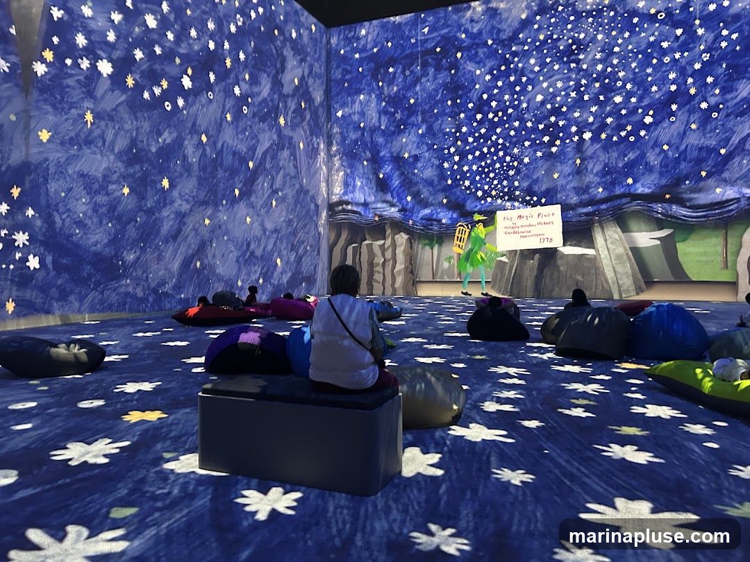 An immersive exhibition space with walls and floor decorated with a starry night sky motif. Several people are seated on beanbag chairs and benches, facing a stage area. On the stage, a figure in green stands next to a sign that reads "The Magic Flute" and lists "Royal Academy of Music" and "Covent Garden Opera 1978".