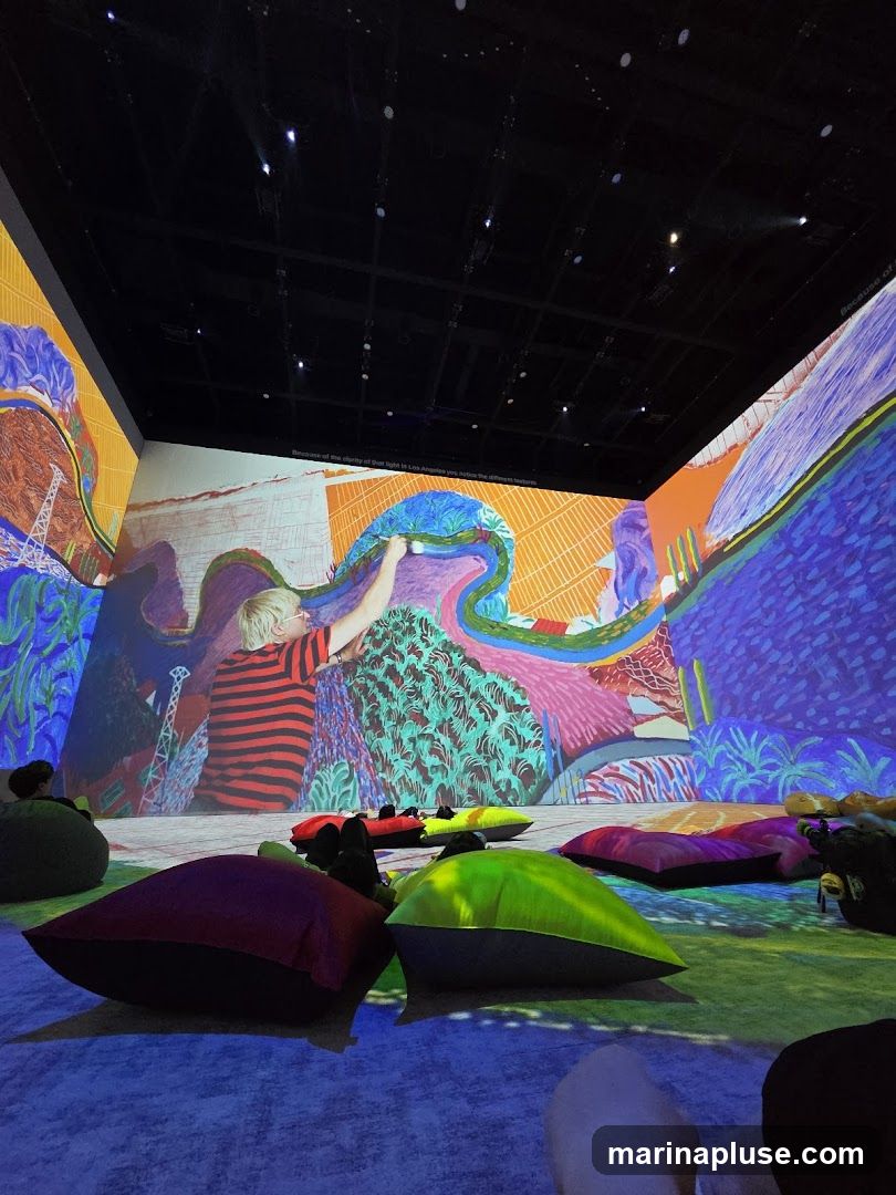 An immersive art installation with vibrant, colorful projections covering the walls and ceiling. A child in a striped shirt is depicted painting on the wall. Beanbag chairs in various colors are scattered on the floor, inviting viewers to relax and enjoy the experience.