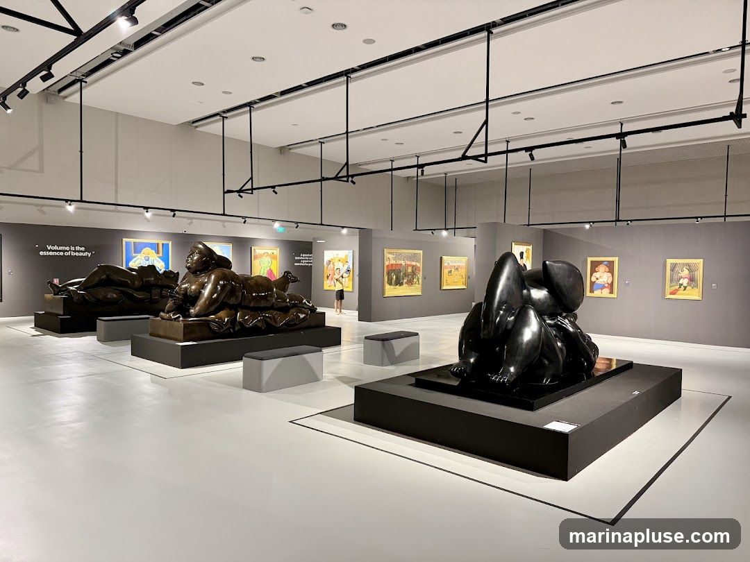 A spacious art gallery with several sculptures and paintings on display. The sculptures are large, dark, and abstract, placed on black pedestals. Paintings with colorful scenes are hung on the grey walls. The lighting is modern and track-based. Benches are provided for visitors.