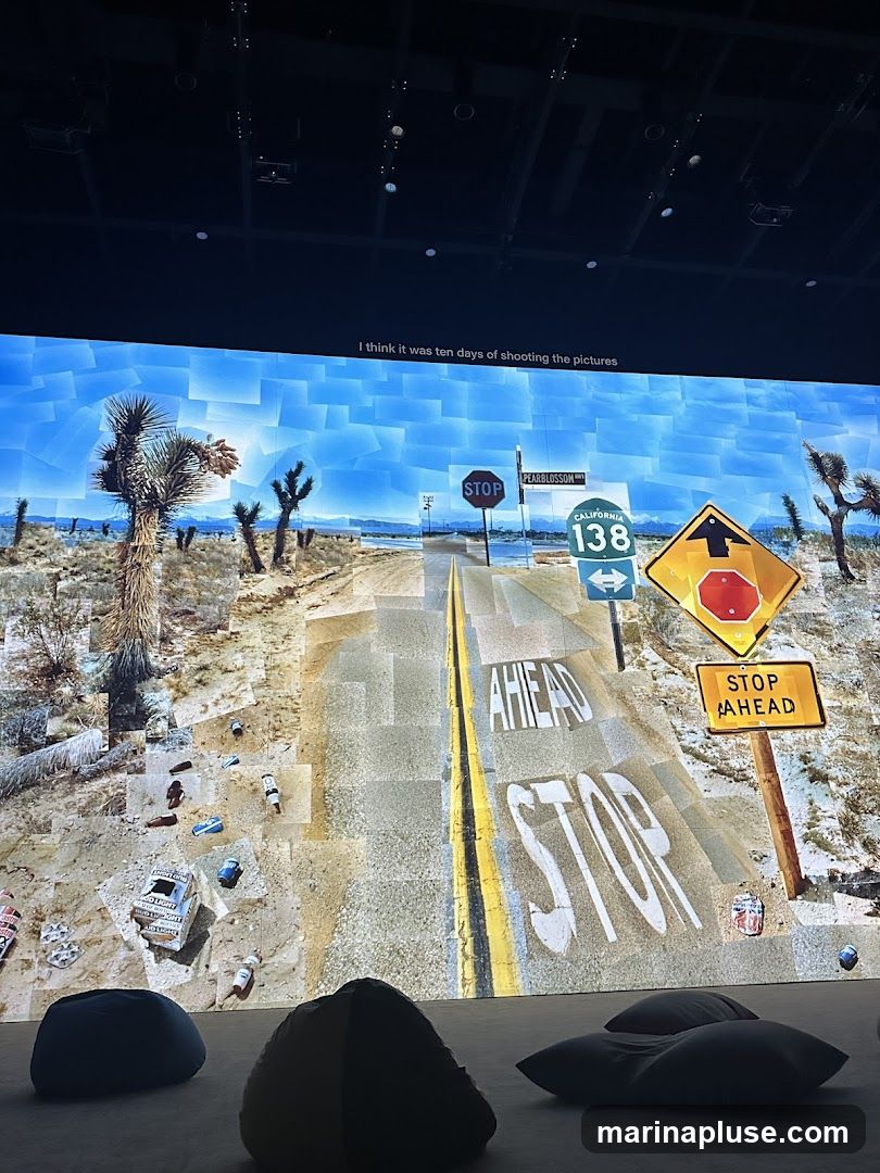 A large screen displays a digital art piece depicting a desert landscape with a road. The road has the words "STOP" and "AHEAD" painted on it. Various road signs, including a stop sign, a California route 138 sign, and a stop ahead sign, are visible. The ground is littered with empty cans and bottles. In the foreground, several large beanbag chairs are placed on the floor. Text at the top of the screen reads, "I think it was ten days of shooting the pictures.".