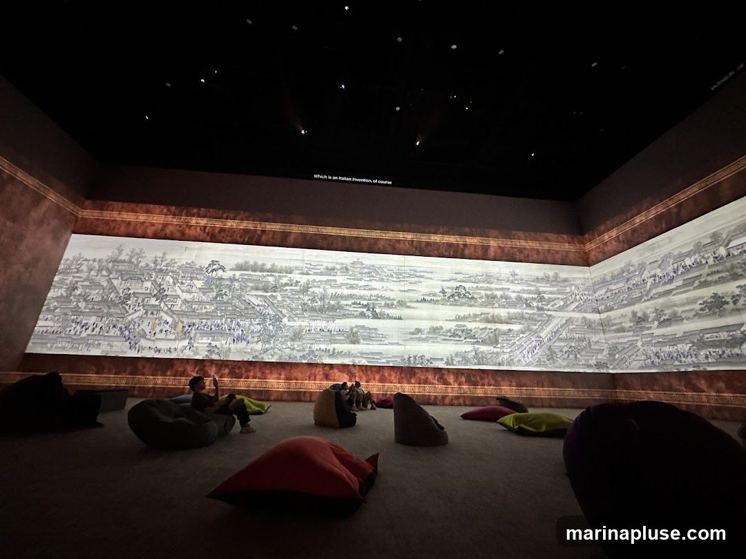 An exhibition hall with a large mural projected onto the wall, depicting a detailed cityscape. Several people are seated on beanbag chairs in the foreground, observing the projection. The room is dimly lit, with spotlights illuminating the artwork. A line of text is visible on the upper part of the wall.