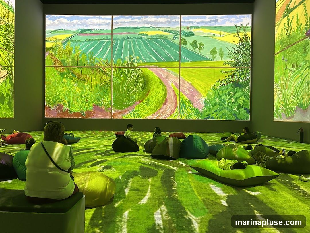 An immersive art installation features large-scale digital projections of a vibrant, sunlit landscape with rolling green fields and a winding path. The room is filled with an assortment of colorful beanbag chairs and floor cushions, where several people are seen relaxing and observing the artwork. The floor is also covered in a green, textured surface that complements the projected imagery.