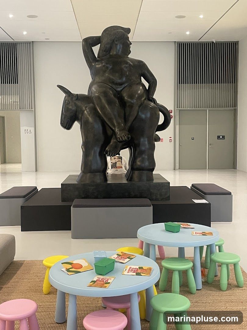 A large bronze sculpture of a woman riding an animal is displayed in a modern, well-lit hall. In the foreground, a children's area features small, colorful tables and stools, with books and small green containers on the tables. Benches are placed around the sculpture.
