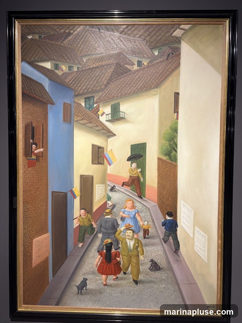 A painting depicts a narrow cobblestone street lined with buildings with red tile roofs. Several people, including children and adults, are walking in the street, some with dogs. Colombian flags are visible on some buildings. There are posters on the walls. The painting is framed in black and gold.
