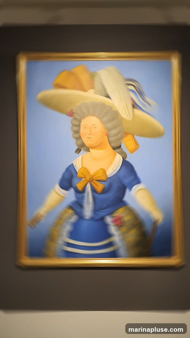 A painting in a gold frame depicts a woman with gray hair styled in a wig. She is wearing a blue dress with a large yellow bow at the neck and a white ruffled collar. Her skirt is wide and patterned. She has a large, wide-brimmed hat adorned with feathers and a yellow ribbon. The background is a solid blue. The painting is slightly blurred.