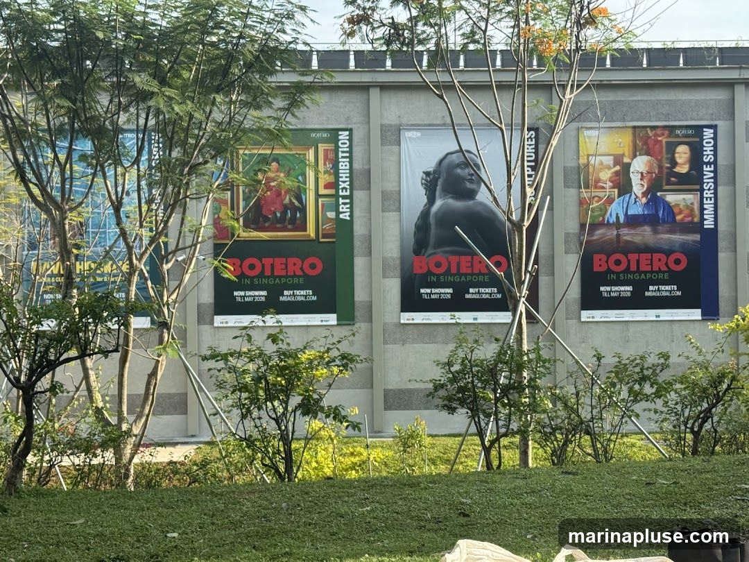 Three large posters advertising a Botero exhibition in Singapore. The posters feature artwork and sculptures by Botero, along with exhibition details such as dates and ticket information. The posters are displayed on a building facade, partially obscured by trees and foliage.